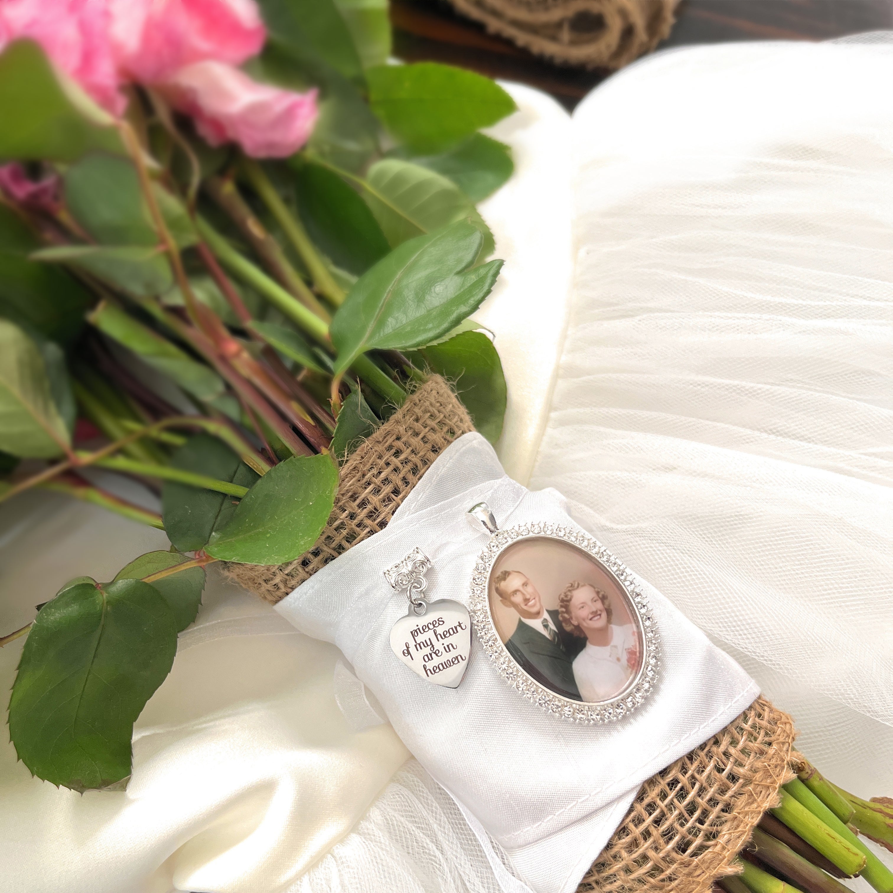 Bridal Memorial Photo Bouquet Charm in oval shape with clear rhinestones around edge of pendant. Image size is roughly 30mmx40mm. Comes with heart shape charm that reads, pieces of my heart are in heaven. Pendant and charm come on a white ribbon to attach to bridal bouquet.