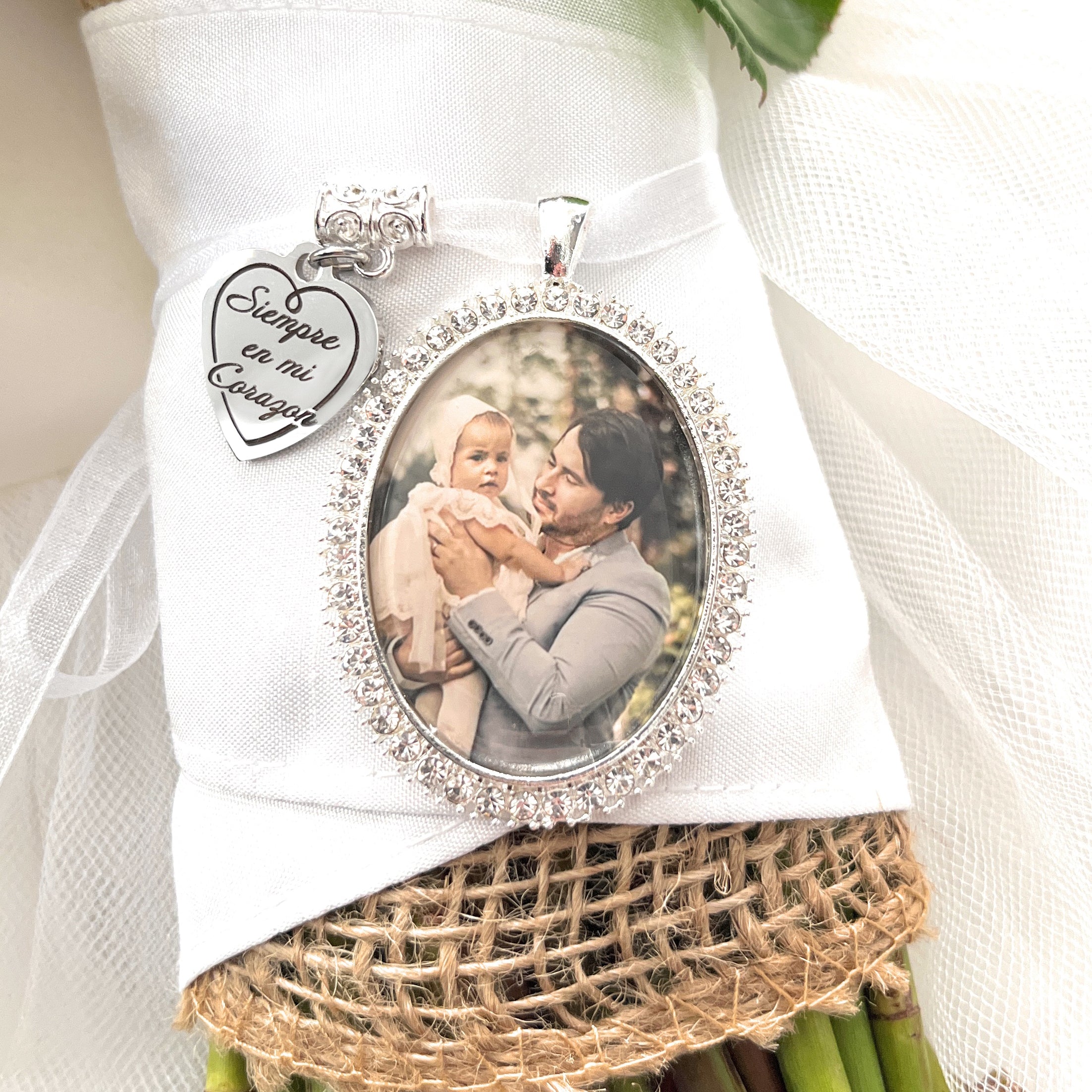 Spanish Bridal Memorial Photo Bouquet Charm in oval shape with clear rhinestones around edge of pendant. Image size is roughly 30mmx40mm. Comes with heart shape charm that reads, Siempre en mi Corazon. Pendant and charm come on a white ribbon to attach to bridal bouquet.