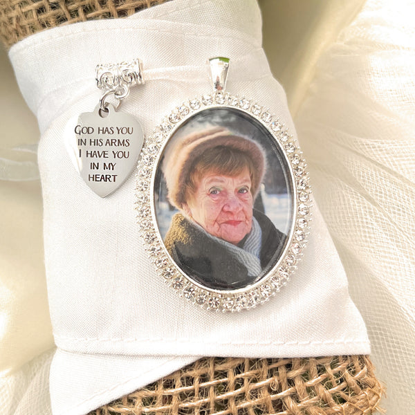 Bridal Memorial Photo Bouquet Charm in oval shape with clear rhinestones around edge of pendant. Image size is roughly 30mmx40mm. Comes with heart shape charm that reads, God has you in his arms I have you in my heart. Pendant and charm come on a white ribbon to attach to bridal bouquet.