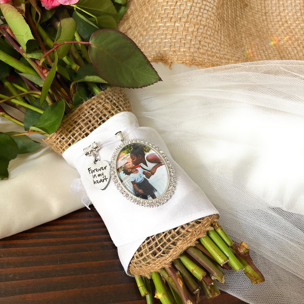 Bridal Memorial Photo Bouquet Charm in oval shape with clear rhinestones around edge of pendant. Image size is roughly 30mmx40mm. Comes with heart shape charm that reads, Forever in my heart. Pendant and charm come on a white ribbon to attach to bridal bouquet.