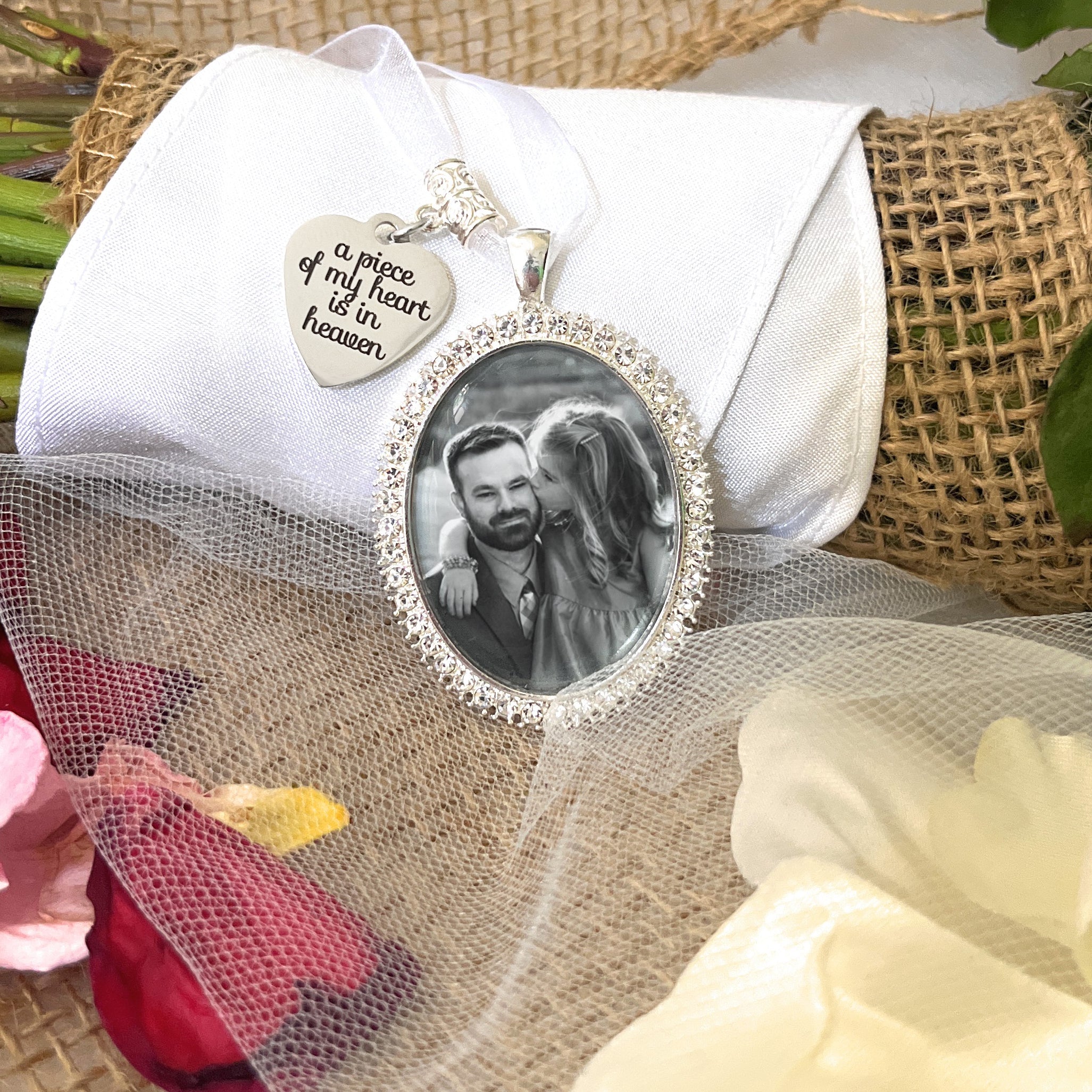 Bridal Memorial Photo Bouquet Charm in oval shape with clear rhinestones around edge of pendant. Image size is roughly 30mmx40mm. Comes with heart shape charm that reads, a piece of my heart is in heaven. Pendant and charm come on a white ribbon to attach to bridal bouquet.